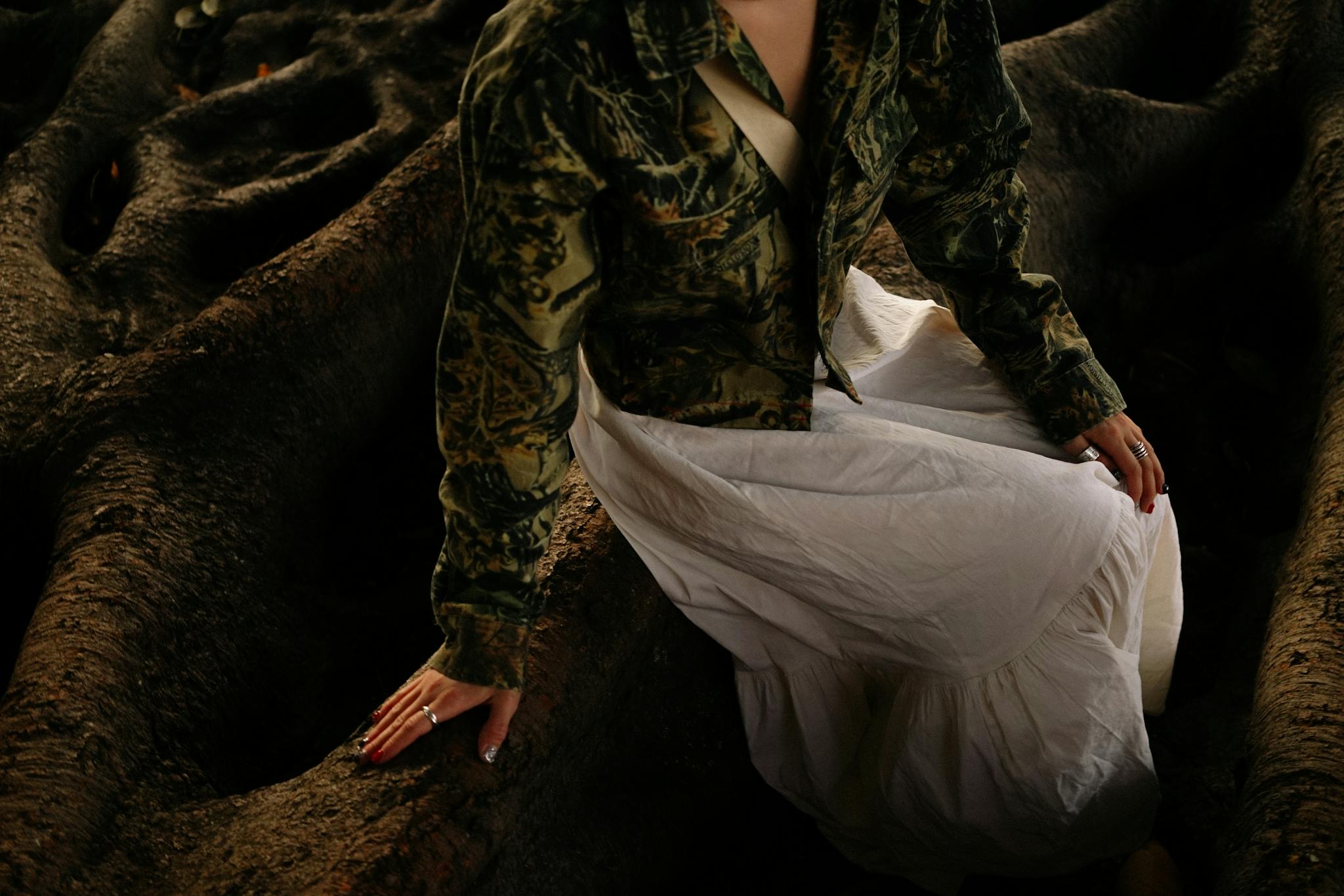 A woman sits on large, textured tree roots wearing a camouflage jacket and white dress.